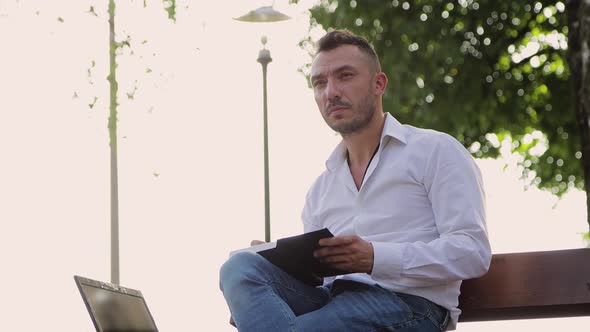 A Concentrated Man in a White Shirt Writes Down Plans in a Notebook on a Bench