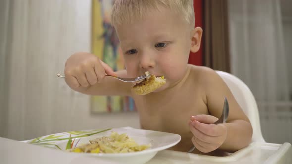 Little cheerful and playful blonde boy learns to eat with a fork on his own.