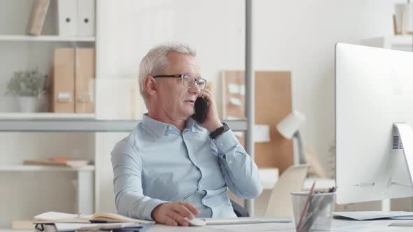 Senior Man Working on Computer and Talking on Smartphone in Office