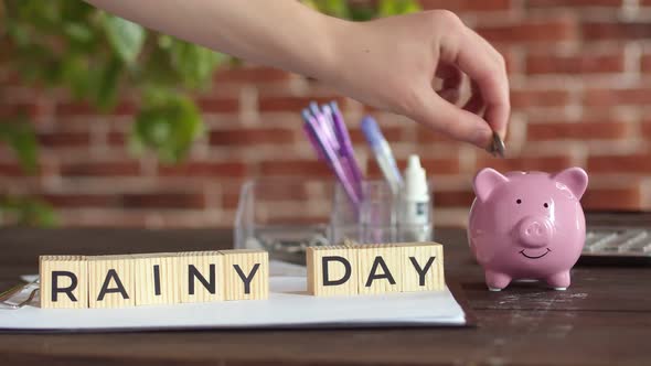 Hand Puts Money in Piggy Bank Next to Which There are Cubes with Inscription Rainy Day