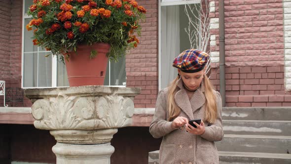 Young Girl in Coat and Beret Using Smartphone on Stairs Brick House in Garden. Beautiful Girl