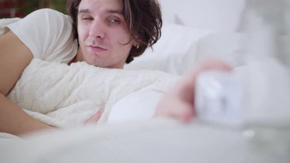 Closeup Portrait of Sleepy Young Man Looking at Alarm Clock and Waking Up with Shocked Facial
