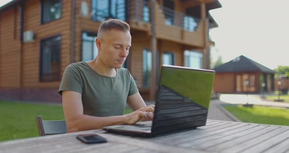 Focused Young Man Working on Laptop While Sitting at Table in Courtyard of Country House