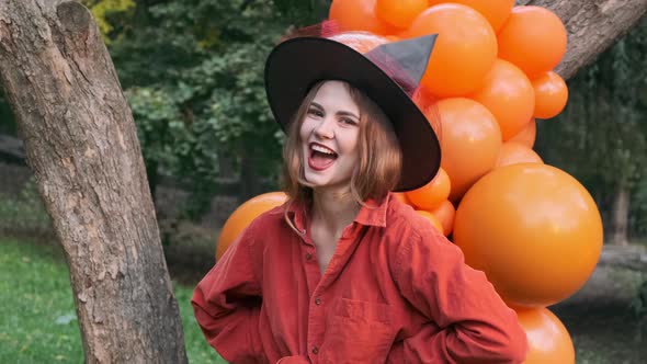 Beautiful girl in an orange shirt, black skirt and witch hat. Posing in front of the camera, laughin