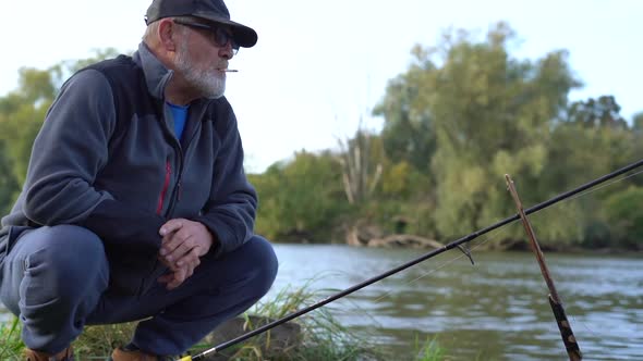 An Elderly Fisherman Sits on His Haunches Near His Spinning Rod and Waits for the Fish to Bite