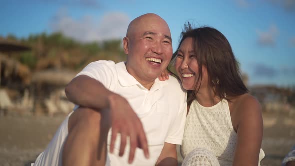 Loving Adult Asian Couple Sitting in Sunlight on Picturesque Mediterranean Sea Laughing Admiring