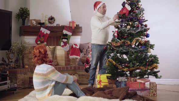 Wide Shot Portrait of Relaxed Caucasian Man in Party Hat Decorating Christmas Tree and Stepping on
