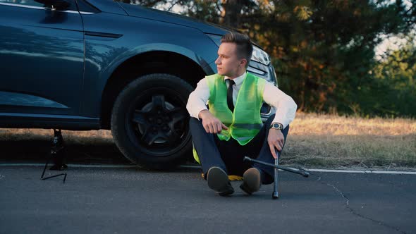 Tired Young Businessman in a Green Safety Vest Has Problems with the Wheel of His Car