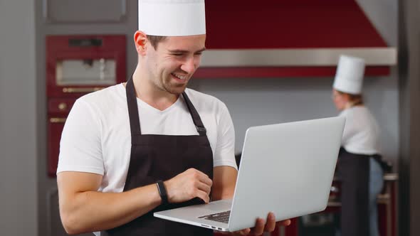 Young Smiling Male Chef in Uniform Standing in Kitchen and Working Online on Laptop