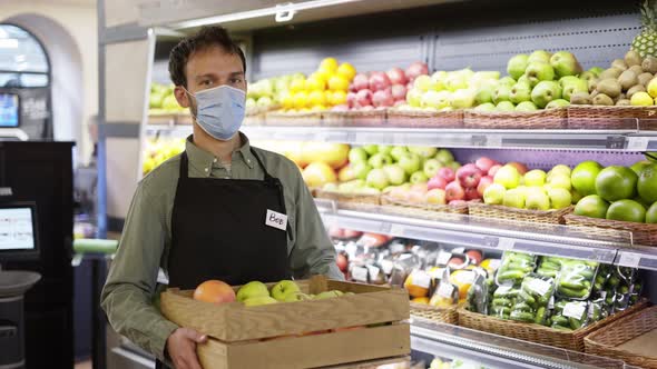 Portrait of Handsome Man in Face Mask and Apron Standing in Food Store with Box of Fruits Indoors