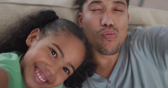 Happy biracial father and daughter sitting on sofa taking selfies