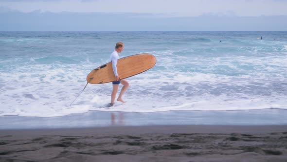 the Surfer Walks Along the Sandy Shore Against the Waves of the Surf