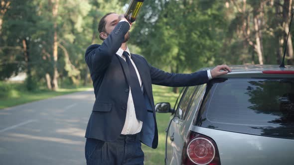 Drunk Young Man Drinking Wine From Bottle Leaning on Car Standing at Suburban Road