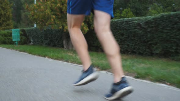 Athletic Handsome Man with Wireless Headphones Jogging Along Sidewalk Near Forest at Summer Day