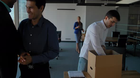 Young Employee Unpack Box with Docs and Equipment. His Colleagues Walking Near in Modern Office