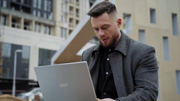 Young businessman types on laptop while sitting on a bench with modern building background.