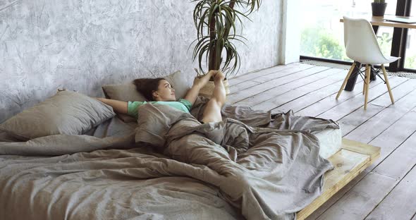 Sleepy Young Woman in Pajamas Wakes Up in Morning at Home, Stretches Leg, Doing Exercise in Cozy Bed