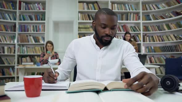 Smart Bearded Dark-Skinned Guy Sitting in the Reading Room on the Bookshelves Background