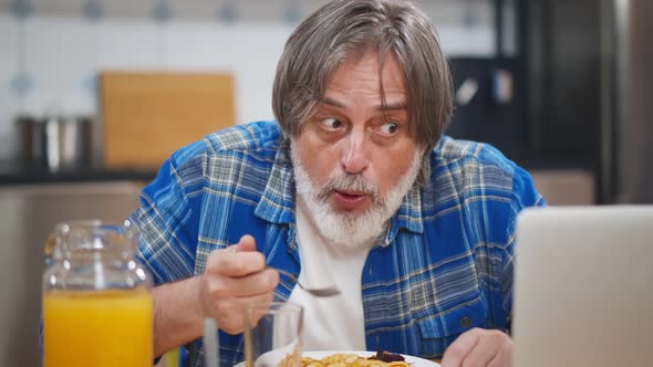 Senior Man with Beard Using Laptop While Having Dinner at Table in Kitchen