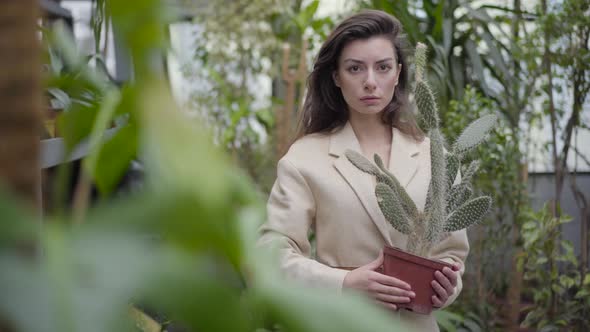 Portrait of Mysterious Caucasian Woman Standing Between Green Plants Holding Pot with Cactus