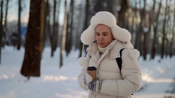 Positive Young Man is Walking Alone in City Park in Winter Holding Hot Drink and Looking at Camera