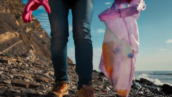 A volunteer in protective gloves collects plastic bottles on a wild beach