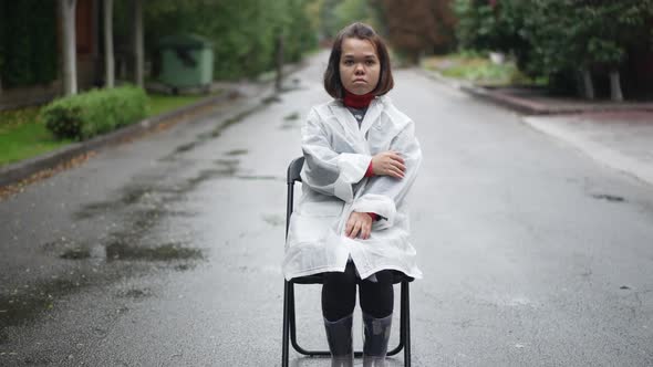 Front View Young Sad Little Woman Looking at Camera Sitting on Chair on Road