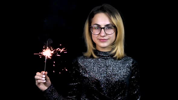 A Girl Holds a Sparkler in Hand. A Young Woman with Glasses Stands in a Shiny Evening Dress