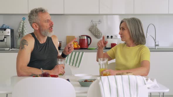 Happy Mature Senior Couple Talking Enjoying Having Breakfast at Home Kitchen