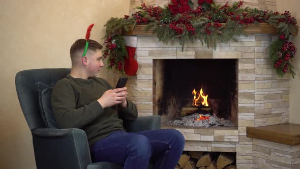 A Young Man Sits in an Armchair By the Fireplace with Horns on His Head and Writes in a Smartphone