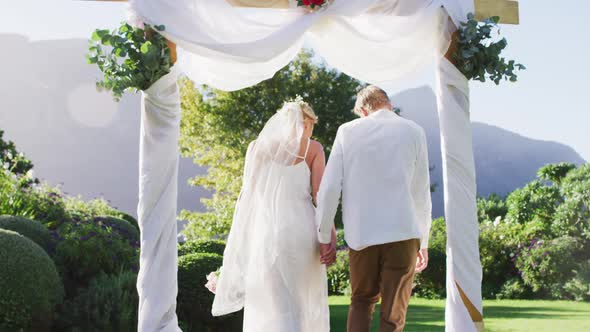 Happy caucasian newly wed couple, holding hands walking from altar outdoors