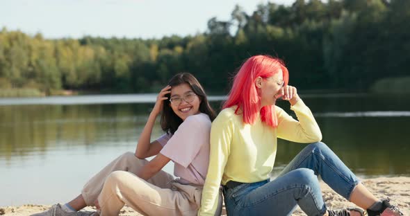 Smiling Girls in Glasses are Sitting on the Sand of the Beach Near the Lake Leaning Against Each