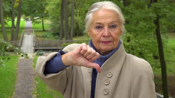 An Elderly Woman Shakes Her Head and Shows a Thumb Down To the Camera in a Park - Closeup