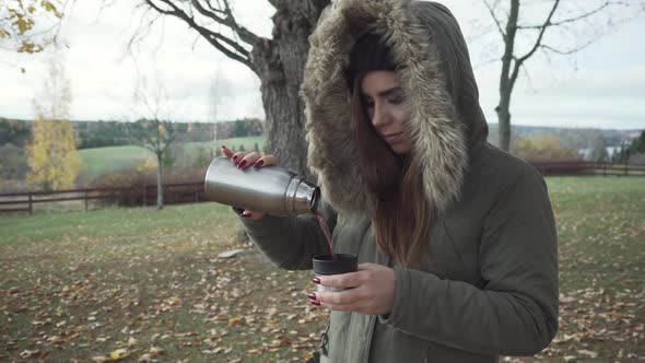 Adult girl is pouring her warm drink and drinking it on cold autumn day.