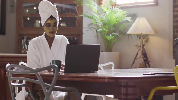 African american attractive woman with applied face mask drinking coffee and using laptop at home