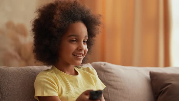 Portrait Little African American Girl Watches TV and Switches Channels with Interest with Remote