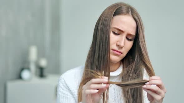 Upset Young Caucasian Woman Looking on Weak Unhealthy Dry Hair Ends at Home Interior