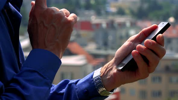Old Senior Man Works (Typing) on Smartphone - City (Buildings) in Background - Closeup