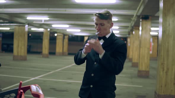 Young Man in a Suit with Bowtie Standing By the Shopping Cart in the Parking at Night and Smoking