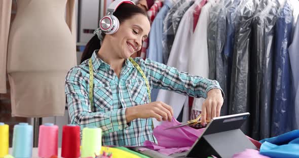 Joyful Woman Seamstress Designer in Headphones Cuts Fabric at Workplace