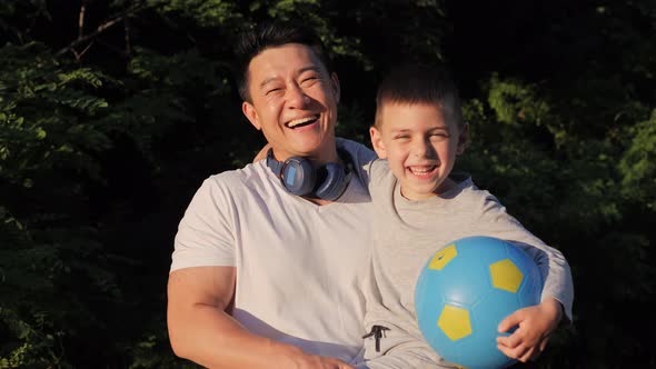 Asian Man in White Tshirt Holding Little Kid Boy with Ball in Arms at Sunset
