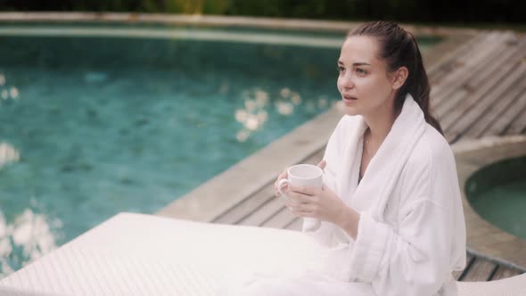 Woman in White Bathrobe Sits on Sunbed Near Pool Drinks Coffee in Early Morning