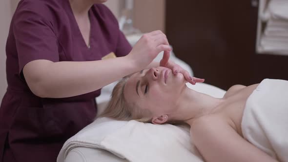 Beautiful Blond Caucasian Woman Enjoying Facial Massage Lying in Spa Salon with Unrecognizable