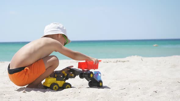 On a Sunny Summer Day, a Boy in Panama Plays with a Toy Excavator on a Sandy Beach, with an Azure