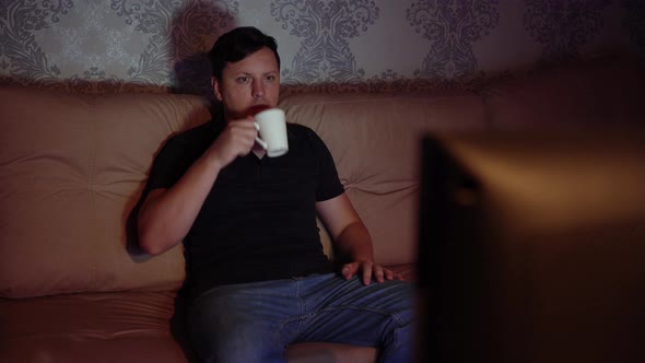 Young Man Watching Tv Sitting on the Couch with a Mug of Tea is Resting at Night
