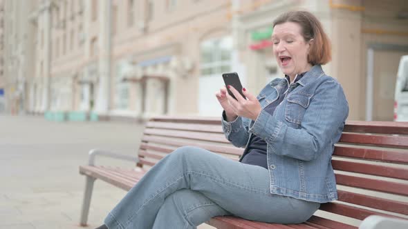 Old Woman Celebrating Online Success on Smartphone While Sitting Outdoor on Bench