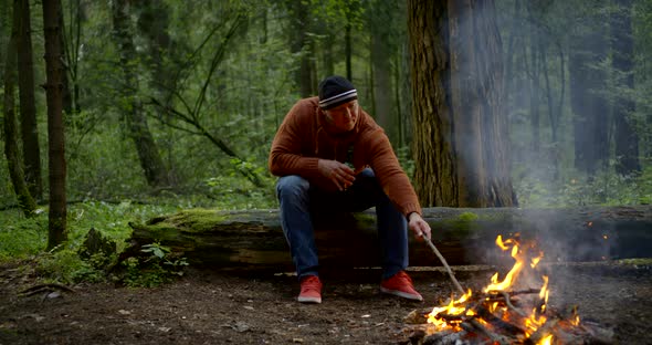 A Middle-aged Man Is in the Woods and He Is Sitting Near a Burning Fire on a Log, a Stick Interferes