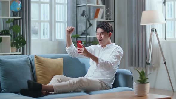 Happy Asian Man Celebrating While Lying On Sofa And Using Smartphone In The Living Room