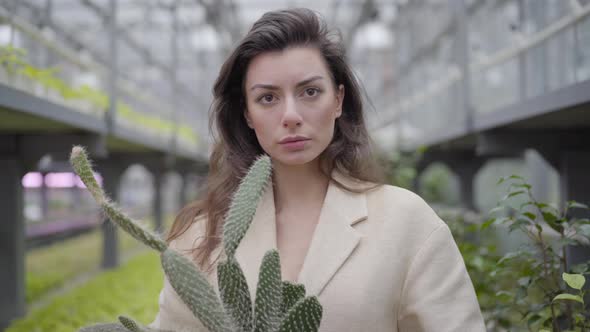 Portrait of Young Brunette Woman in Elegant Beige Jacket Holding Cactus and Looking at Camera