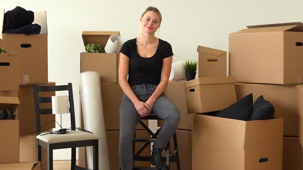 A Friendly Moving Woman Sits on a Chair in an Empty Apartment and Waves at the Camera with a Smile